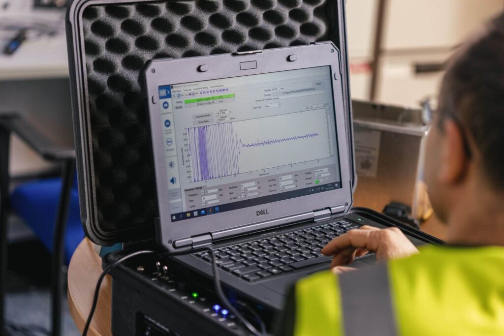 Man in a high visibility yellow jacket. He has a DELL laptop open and facing him in a hard case. On the screen, he is measuring waves from an object for NDT Research and Development purposes.